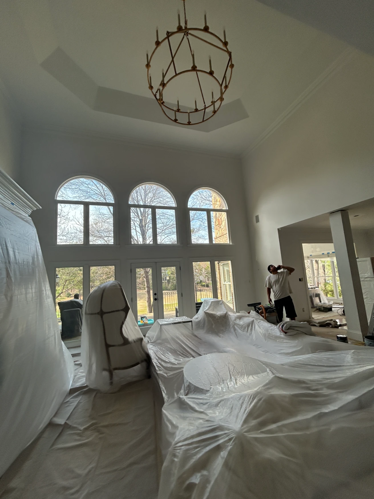 Living room during remodeling, furniture covered in plastic, high ceilings, large windows, chandelier